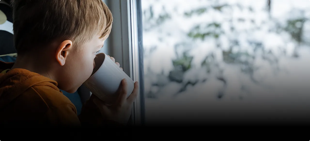 A child drinking from a mug beside a frosted window on a winter day