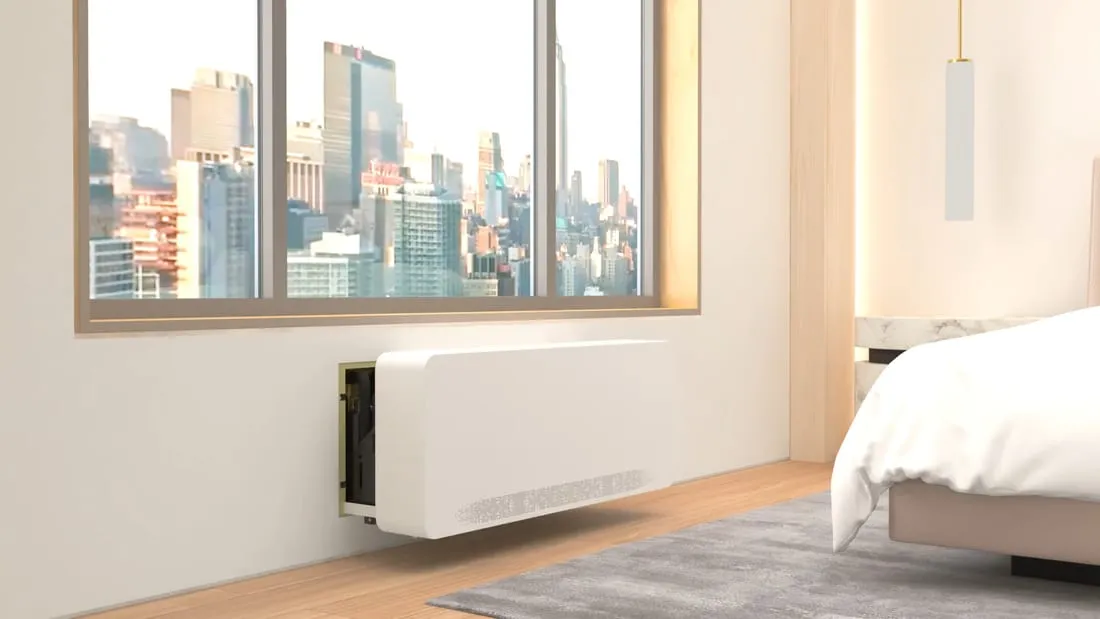 A white through-wall unit under a hotel-room window with a city view, slid partway out of its wall sleeve